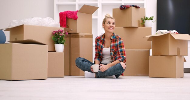 people, moving new place and repair concept   happy beautiful  young woman with many cardboard boxes sitting on floor with Cup at home