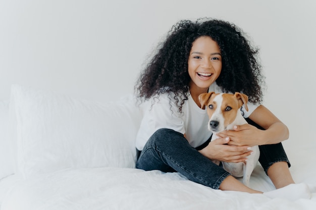 People, pet care and friendship concept. Smiling Afro woman with glad expression cuddles pedigree dog, sit on comfortable bed