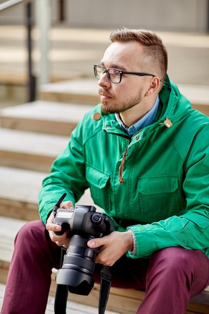 people, photography, technology, leisure and lifestyle - young hipster man holding digital camera with big lens and sitting on stairs in city