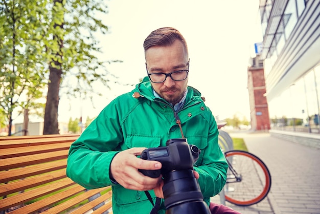 people, photography, technology, leisure and lifestyle - young hipster man holding and looking to digital camera with big lens on city street