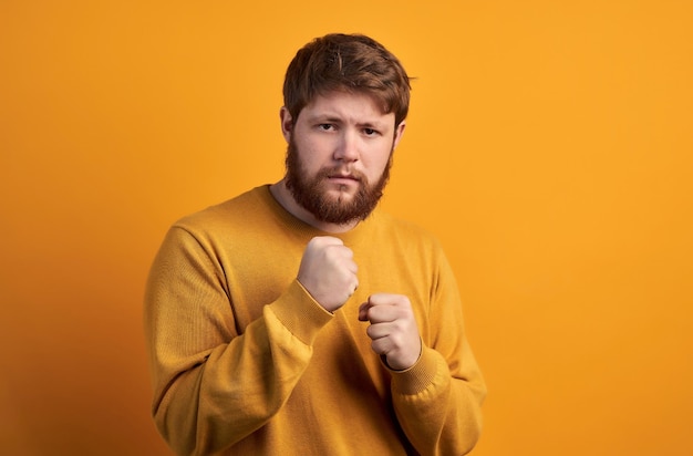 People and power concept Serious foxy man with determined expression keeps clenched fists looks with stern at camera ready to fight dressed in dark blue shirt stands against white wall