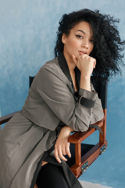 People, race, ethnicity and fashion concept - beauty black model posing in high boots. Young pretty beautiful mulatto woman sitting in director's chair at film. woman in gray raincoat, afro hairstyle