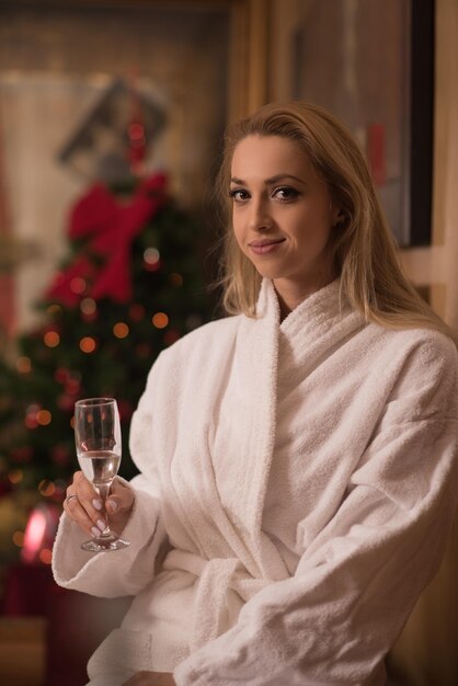 people, and relaxation concept   beautiful young woman in bath robe drinking champagne at spa over holidays lights background