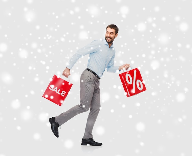 people, sale, christmas, winter and holidays concept - smiling man holding red shopping bags with percentage sign over snow background