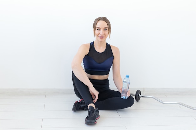 People, sport and fitness concept - young woman sitting with bottle of water on gym mat.