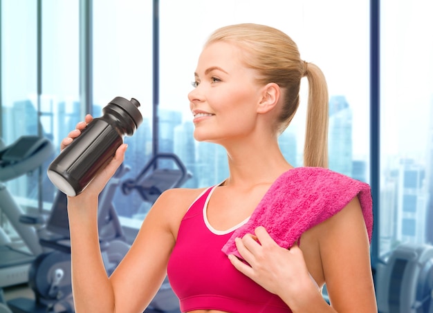 people, sport, fitness and recreation concept - happy woman drinking water from bottle with towel over gym machines background