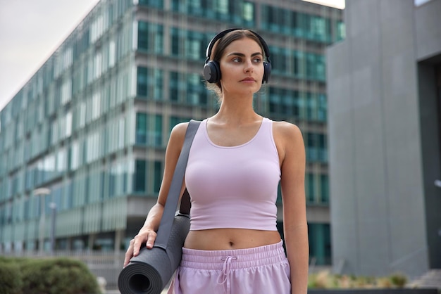 People sport and recreation concept. Thoughtful sportive woman dressed in activewear strolls in urban setting listens music via headphones carries rubber rolled kareamt leads healthy lifestyle