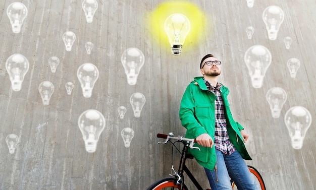 people, startup, inspiration and lifestyle - happy young hipster man with fixed gear bike on city street over concrete wall and lighting bulbs background