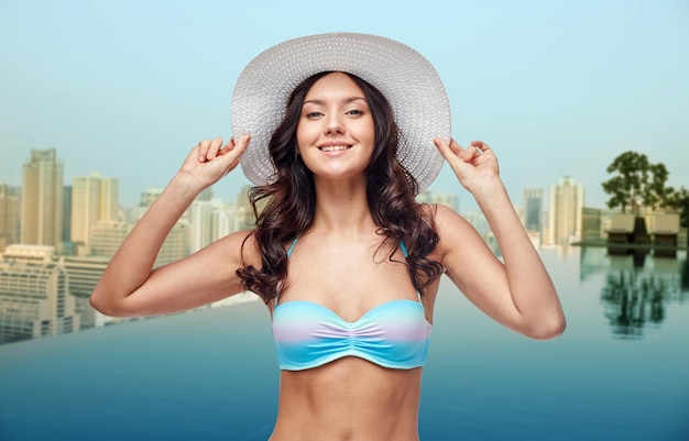 people, summer holidays, travel and tourism concept - happy young woman in bikini swimsuit and sun hat over infinity edge swimming pool and city background