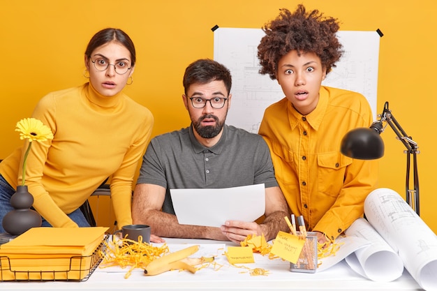 People teamwork and collaboration concept. Three shocked collaegues partners cooperate for creating architectural plan stare surprised, pose at office desk work on building project.