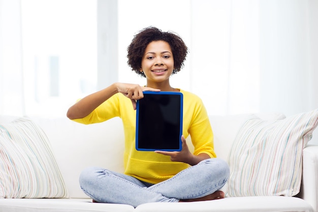 people, technology, advertisement and leisure concept - happy african american young woman sitting on sofa and showing tablet pc computer black blank screen at home