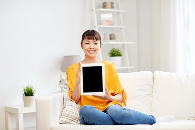 people, technology, advertisement and leisure concept - happy young asian woman sitting on sofa and showing tablet pc computer blank black screen at home