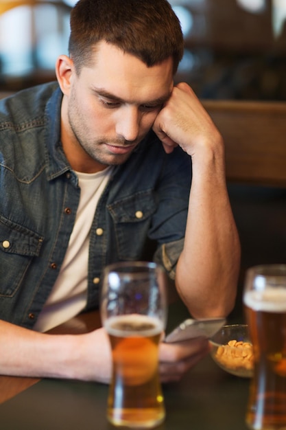 people and technology concept - man with smartphone drinking beer and reading message at bar