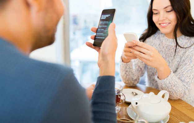 people, technology and dating concept - happy couple with smartphones drinking tea at cafe or restaurant