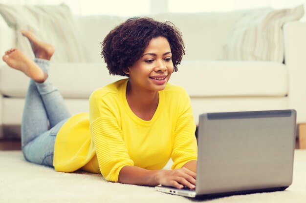 people, technology and leisure concept - happy african american young woman lying on floor with laptop computer at home