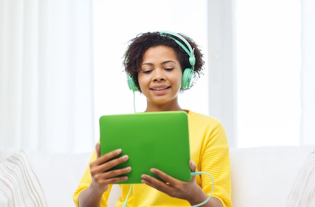 people, technology and leisure concept - happy african american young woman sitting on sofa with tablet pc computer and headphones listening to music at home