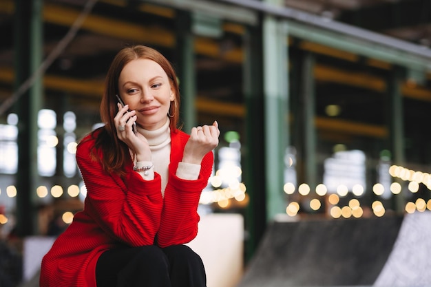 People technology and lifestyle concept Positive Caucasian woman has telephone conversation talks to friends via cellular wears red coat and black trousers poses outside with free space