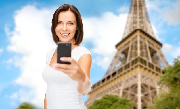 people, technology, tourism and travel concept - young woman taking selfie with smartphone over paris eiffel tower background