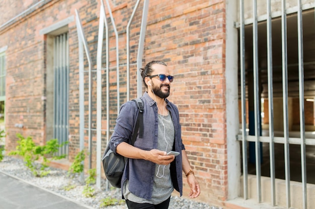 people, technology, travel and tourism - man with earphones, smartphone and bag walking along city street and listening to music