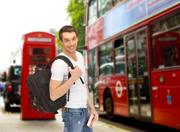 people, travel, tourism and education concept - happy young man with backpack and book over london city bus on street background