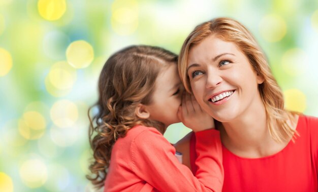 people, trust, love, family and motherhood concept - happy daughter whispering gossip to her mother over green lights background