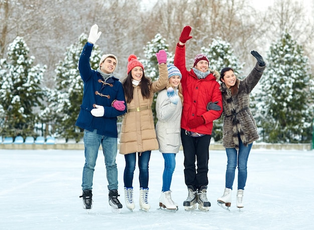 people, winter, friendship, sport and leisure concept - happy friends ice skating and waving hands on rink outdoors