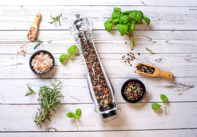 Pepper mill with different peppers in bowl with himalayan salt and fresh rosemary and basil on the wooden background