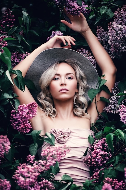 Perfect Blonde Model in Spring Flowers Garden. Beautiful Woman with Healthy Skin, Beauty Hairstyle and Fashionable Hat