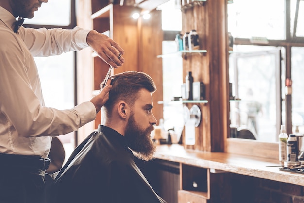 Perfect cut. Close-up side view of young bearded man getting haircut by hairdresser at barbershop