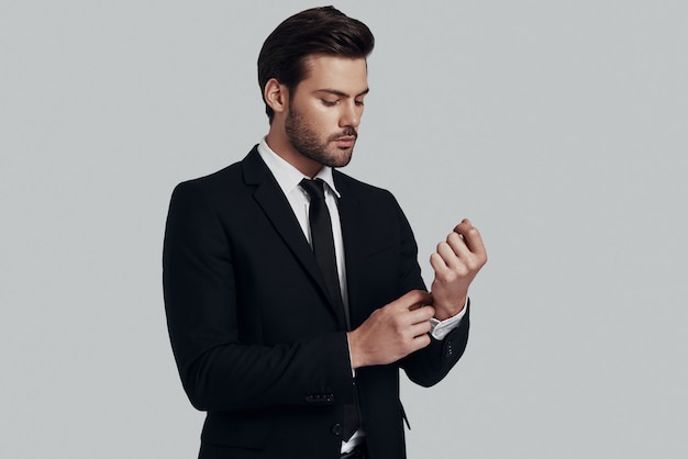 Perfect in every way. Handsome young man in full suit adjusting sleeve while standing against grey background