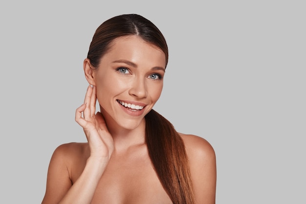 Perfect skin. Attractive young woman looking at camera and smiling while standing against grey background