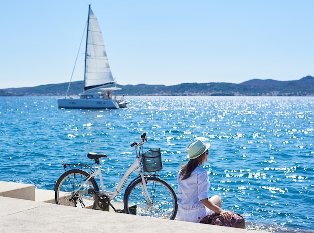 Perfect summer landscape on bright sunny day. Back view of female tourist with backpack sitting at bicycle on paved sidewalk under clear blue sky. Sailing ship in azure water