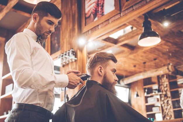 Perfect trim at barbershop. Low angle view of young bearded man getting haircut by hairdresser with electric razor while sitting in chair at barbershop