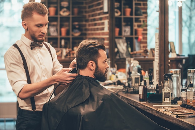 Perfect trim at barbershop. Young bearded man getting haircut by hairdresser with electric razor 