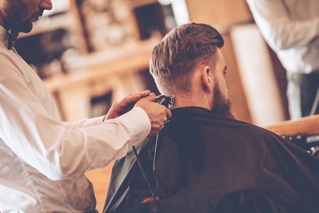 Perfect  trim. Rear view close-up of young bearded man getting haircut by hairdresser with electric razor while sitting in chair at barbershop