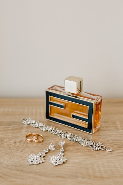 A perfume bottle, a bracelet, earrings-studs and wedding rings on a wooden table against a light wall