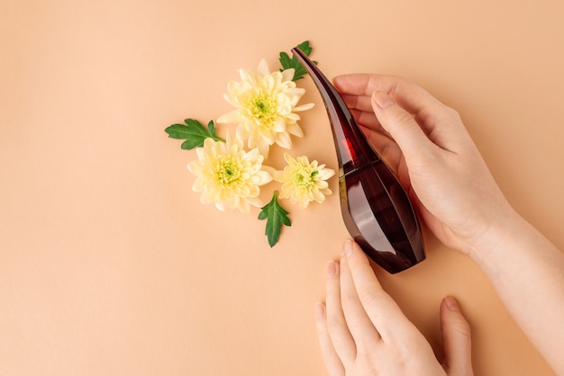 Perfume in female hands, flowers and green leaves on beige