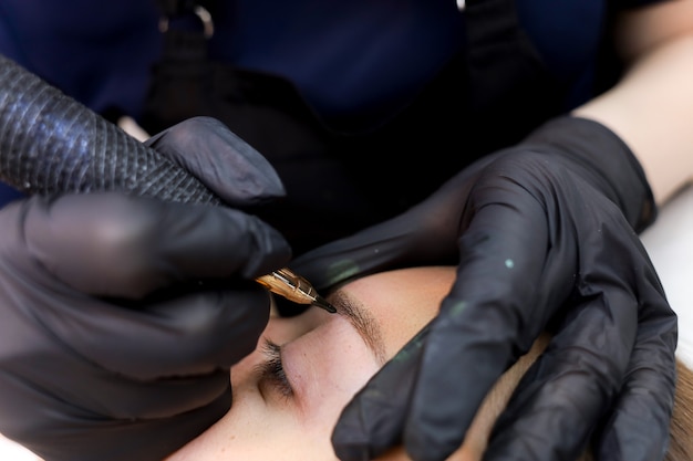 The permanent makeup artist leans over the client and performs an eyebrow tattoo