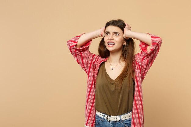 Perplexed shocked young woman in casual clothes looking aside, putting hands on head isolated on pastel beige wall background in studio. People sincere emotions, lifestyle concept. Mock up copy space.