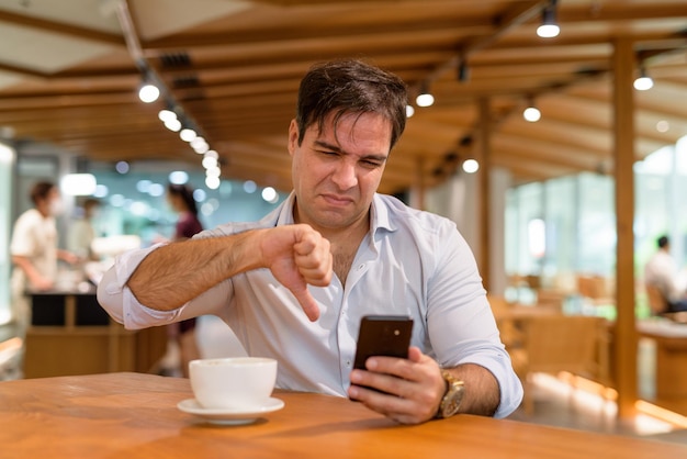 Persian man sitting at coffee shop while using mobile phone and getting bad news