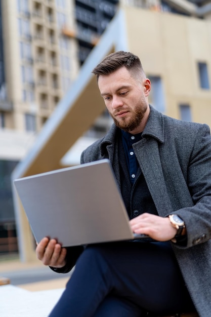 Persistent businessman in gray casual coat doing work on laptop Modern wide street on background Business concept