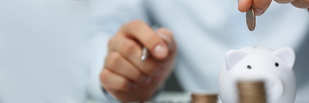 Person accountant put coin in white piggy bank, stack of coins on workplace