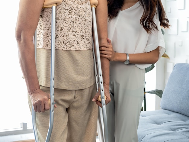 Person helping female's hand hold the arms of an elderly woman trying to walk on crutches standing in recovery room background Helping and care senior patient to walk with crutches walker concept