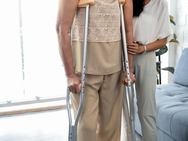 Person helping female's hand hold the arms of an elderly woman trying to walk on crutches standing in recovery room background Helping and care senior patient to walk with crutches walker concept