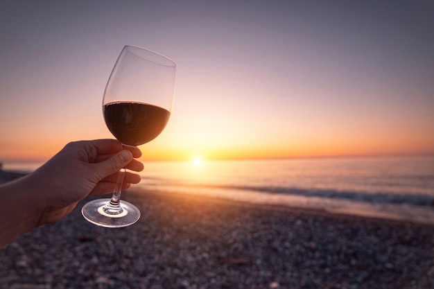 Person holding glass of red wine during watching sunset and enjoying sea holiday