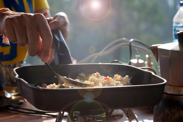 A person is cooking fried rice in a pan with blur green field background at a morning