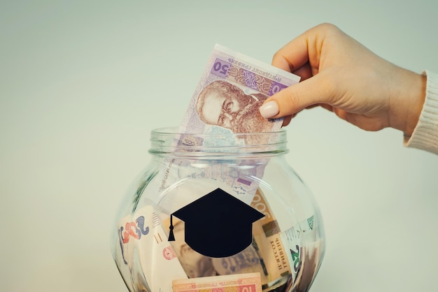 Person putting hryvnia banknote to the glass jar with students hat symbol Saving money for education in Ukraine toned photo