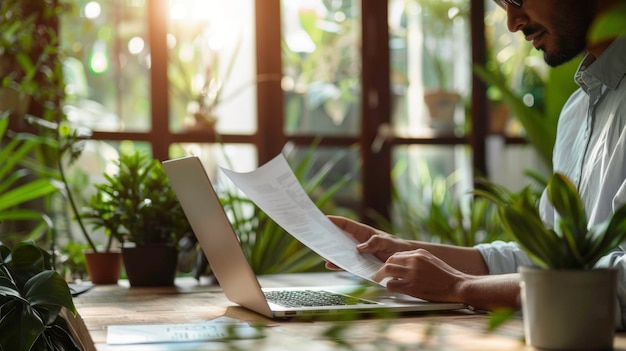 A person reviews documents while working on a laptop in a bright plantfilled workspace that promotes