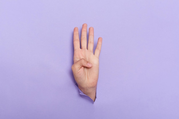 Person's hand sticks out of hole in paper of lilac background four fingers sign copy space for advertisement