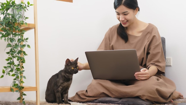 Pet lover concept Young Asian woman using laptop and playing to stroking with cat in living room